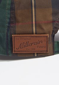 Brązowa skórzana metka na kratkowanej tkaninie, z zielonymi, ciemnoniebieskimi i brązowymi paskami. Tekst zawiera "Millerain Fabric Est. 1880."
