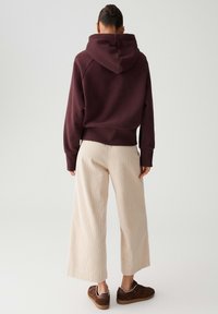 Persona con i capelli raccolti, che indossa un felpa cappuccio marrone scuro, pantaloni ampi color crema con trama e sneakers marroni, in piedi di fronte a uno sfondo semplice.