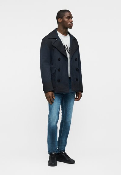 Homme portant un manteau croisé bleu marine par-dessus un t-shirt blanc à motif, un jean bleu et des baskets noires, debout, tourné vers la droite.