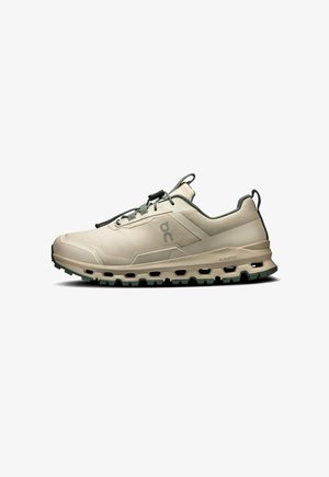 Light beige running shoe with a mesh upper, green accents, and a textured sole featuring deep treads. Minimalist design with elastic laces.