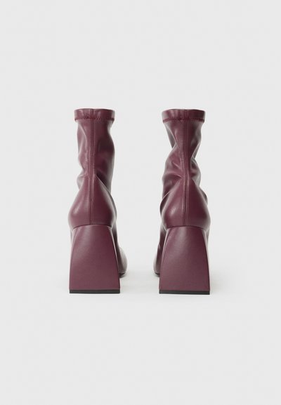 Bottes de cheville en cuir bourgogne avec un design loose, des talons carrés et des détails de couture visibles. Texture lisse, silhouette minimaliste.