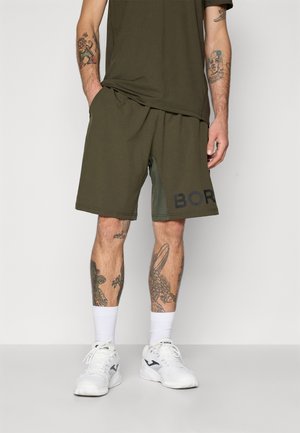 Olivengrønne sports shorts med sort "BOR" tekst, lavet af blødt stof, parret med hvide sokker og sko. Tatoveringer synlige på benene.