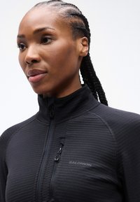 Γυναίκα με πλεξούδες που φοράει μαύρο μπουφάν Salomon με φερμουάρ και τσέπη στο στήθος, πάνω σε απλό λευκό φόντο.