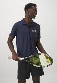 Navy polo shirt with short sleeves and a collar, featuring a white EA7 logo. The model holds a black and green tennis racket with a ball.
