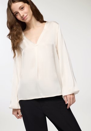 Blusa a maniche lunghe color crema chiaro con scollatura a V, texture liscia e vestibilità rilassata, abbinata a pantaloni neri. Design pulito e minimalista.