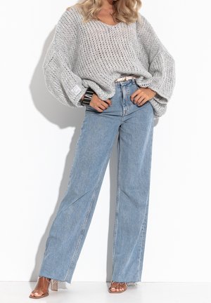 Femme portant un jean bleu clair ample, un pull en maille gris oversize, un large bracelet rayé et des sandales à talons hauts ouvertes transparentes.