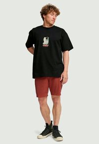 Young man wearing black graphic t-shirt, rust-colored shorts, and black high-top sneakers standing against plain background.