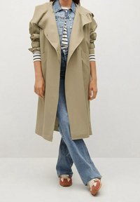 Trench-coat beige avec un large col, porté sur une veste en jean et une chemise rayée. Jean évasé bleu et baskets bicolores complètent la tenue.