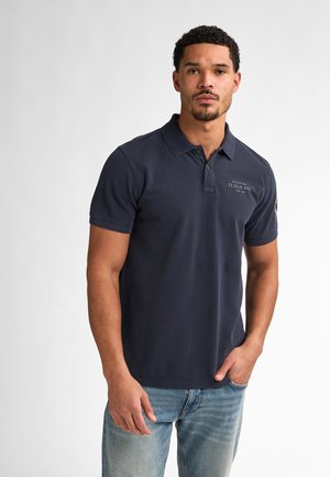 Navy blue polo shirt with short sleeves, soft fabric, three-button placket, and subtle logo detailing on the chest. Worn with light blue jeans.