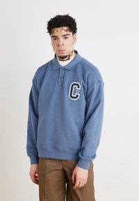 Pull bleu en coton avec col et manches longues. Présente un logo brodé blanc sur la poitrine gauche. Texture lisse, coupe décontractée.