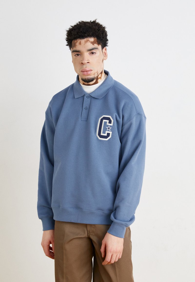 Pull bleu en coton avec col et manches longues. Présente un logo brodé blanc sur la poitrine gauche. Texture lisse, coupe décontractée.