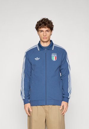 Kék cipzáras dzseki fehér háromcsíkos díszítéssel, bordás mandzsettákkal és szegéllyel. Bal mellrészen hímzett Olaszország címerrel. Simább textúrájú.