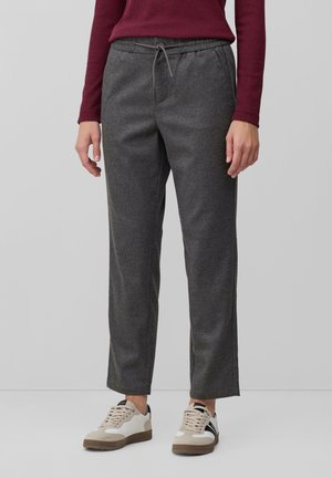 Person iført mørkegrå snor-bukser, mørkerød langærmet top og hvide sneakers med beige og sorte detaljer, stående på en grå gulv.