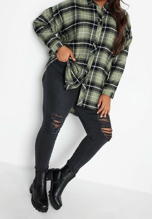 Kvinde iført oversize grøn ternet skjorte, sorte, revnede skinny jeans og sorte chunky ankelstøvler, poserer med hånden i lommen.