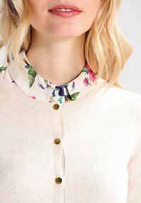 Bežový pletený cardigan s kulatým výstřihem a zlatými knoflíky, překrytý květinovou blůzou s výraznými zelenými a růžovými akcenty.