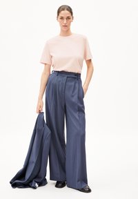 Lichtroze katoenen t-shirt gecombineerd met hooggetailleerde, marineblauwe wijde broek. Het model houdt een bijpassende blazer vast en draagt zwarte schoenen.