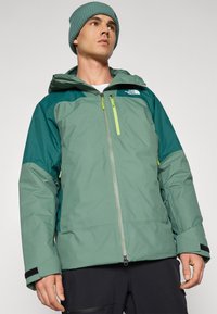 Veste d'hiver verte et teal avec une fermeture éclair à l'avant, une capuche et des poignets ajustables ; comprend un logo sur la poitrine et des poches zippées.