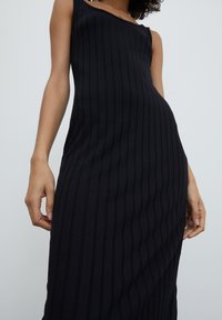 Aansluitende mouwloze jurk, zwarte ribstof, verticale strepen, getailleerde silhoutte, zachte textuur, licht gebogen decolleté, middellange lengte.