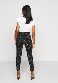 Calças pretas de bolinhas com um corte slim, com bolinhas brancas, combinadas com uma blusa branca de mangas curtas e sandálias de salto. Vista traseira.