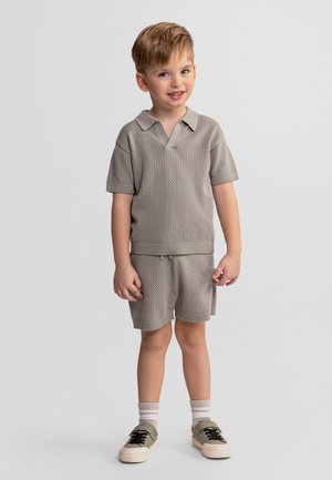 Jonge jongen staand, gekleed in een bijpassend beige textuur korte mouwen shirt en korte broek, witte sokken en beige sneakers met zwarte veters.