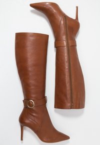 Bottes en cuir marron montant jusqu'aux genoux, avec un bout pointu, un talon stiletto haut, une fermeture éclair latérale et un détail de boucle circulaire sur la sangle de la cheville.