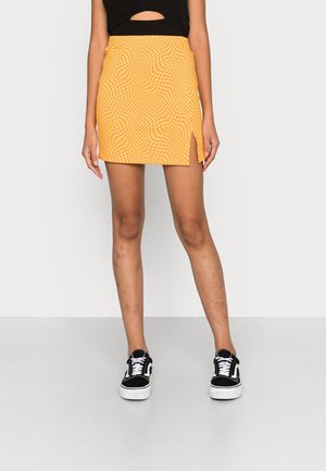 Eine Person steht in einem schwarzen ärmellosen Top und einem gemusterten gelben Minikleid mit einem Seitenschlitz, ergänzt durch schwarze und weiße Sneakers.