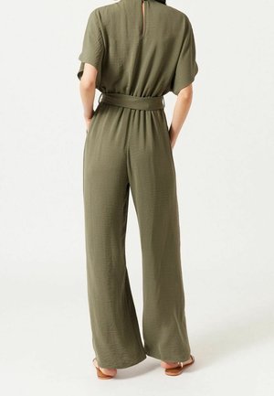 Oliwkowy kombinezon o szerokich nogawkach, krótkich rękawach i pasku w talii. Teksturowana tkanina i detal w kształcie dziurki na plecach.