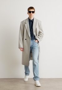 Cappotto di lana grigio chiaro con revers oversize, camicia nera a fantasia, jeans azzurri chiari e sneakers bianche, accompagnati da occhiali da sole neri.