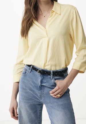Femme portant un chemisier à manches longues jaune pâle rentré dans un jean bleu clair avec une ceinture noire cloutée et une bague en or à la main droite.