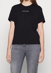 Kvinde iført en sort t-shirt med teksten "MANKIND" i hvid, parret med slidte grå jeans, stående foran en ensfarvet baggrund.