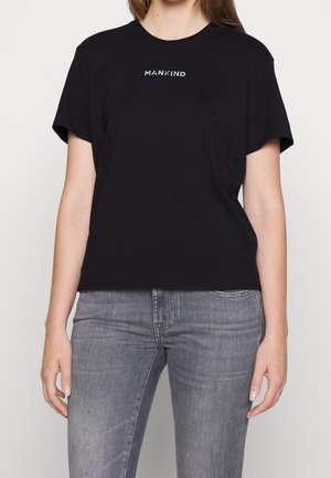 Femme portant un t-shirt noir avec le texte "MANKIND" en blanc, associé à un jean gris délavé, se tenant devant un fond uni.