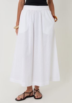 Kobieta ubrana w białą spódnicę o wysokim stanie, sięgającą kostek, z kieszeniami, czarne sandały na paski, czarny top oraz masywne bransoletki.