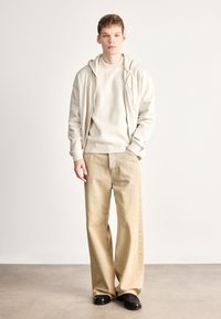 Férfi ruházat világos bézs zipzáras kapucnis pulóverrel, bordázott krémszínű pulóverrel és széles szárú, fakó középső varrással ellátott bézs nadrággal.