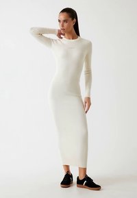 Robe en tricot côtelé crème avec manches longues, col rond et coupe ajustée, associée à des baskets noires à semelle gomme.