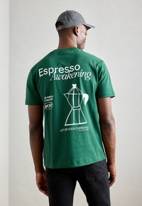 Green cotton t-shirt featuring white text: "Espresso Awakening" and a coffee maker graphic. Short sleeves and relaxed fit.