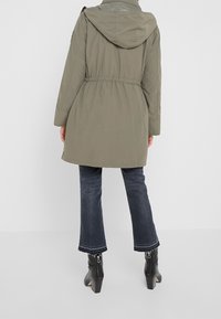 Person som bär en olivgrön huva, mörka korta jeans med fransiga muddar och svarta ankelstövlar med klack, står framför en enfärgad bakgrund.