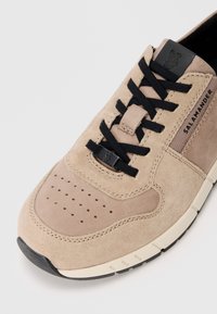 Beige Wildlederschuh mit schwarzen Schnürsenkeln, perforierter Zehenpartie und seitlicher Markenkennzeichnung. Verfügt über eine strukturierte Gummisohle und dezente kontrastierende Akzente.