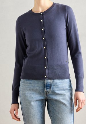 Donna che indossa un cardigan con bottoni a maniche lunghe blu navy e jeans di denim chiaro, in piedi davanti a uno sfondo semplice.
