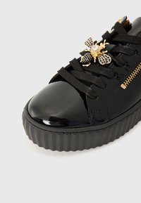 Sneaker in pelle lucida nera con suola rigata, caratterizzato da decorativi embellishments a forma di ape e dettagli in oro, inclusa una zip.