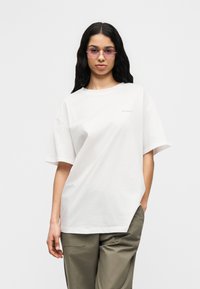 Γυναίκα με μακριά σκούρα μαλλιά που φοράει ανοιχτόμωβ γυαλιά ηλίου, λευκό φαρδύ μπλουζάκι και ελιά πράσινο παντελόνι σε απλό φόντο.