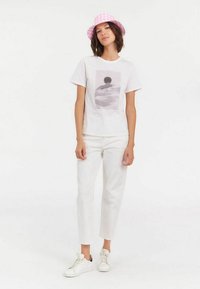 T-shirt blanche avec un imprimé graphique, portée avec un pantalon blanc et un chapeau de pêcheur à carreaux rose. Les chaussures sont des baskets blanches.