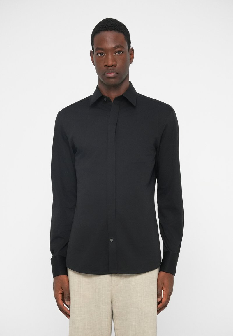 Μαύρη μακριμάνικη μπλούζα από μαλακό ύφασμα με μυτερό γιακά και κουμπιά μπροστά. Διαθέτει στενή γραμμή και κουμπωτές μανσέτες.