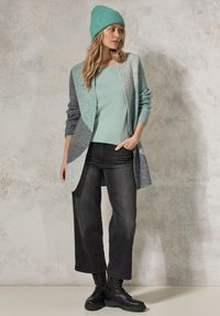 Lichtblauwe en grijs gemêleerde cardigan met een afgeronde, open voorkant, gecombineerd met een muntkleurig topje, zwarte wijde jeans en zwarte veterschoenen.