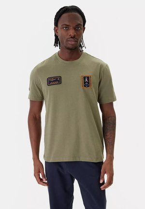 Uomo con capelli intrecciati che indossa una maglietta verde oliva con due toppe sul petto e pantaloni blu scuro, in piedi davanti a uno sfondo bianco.