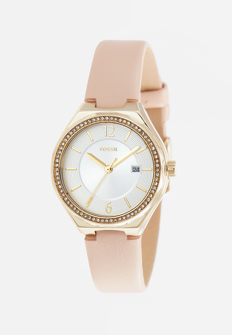 Montre Fossil avec un boîtier doré et un cadran crème. Elle comporte une lunette ornée de cristaux, des index horaires dorés et un bracelet en cuir rose.