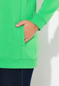 Felpa verde realizzata in morbido tessuto, con una tasca laterale e polsini a coste. Il design è aderente con una texture liscia.