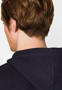 Granatowy hoodie z teksturowanej tkaniny. Posiada kaptur, widoczne szwy oraz widok z tyłu z bliska, ukazujący długość i styl włosów.