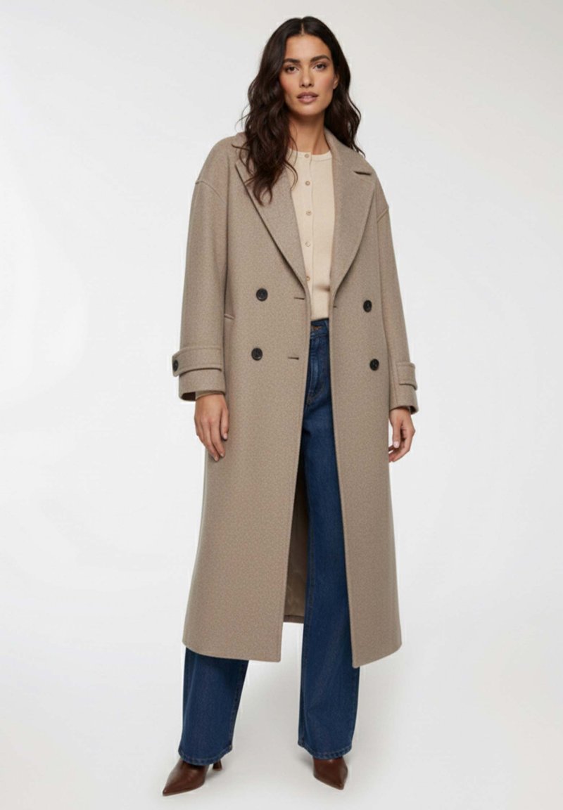 Kobieta z falującymi, ciemnymi włosami, ubrana w beżowy, długi, podwójnie zapinany płaszcz, kremową bluzkę z guzikami, niebieskie spodnie typu wide-leg oraz brązowe, spiczaste buty.