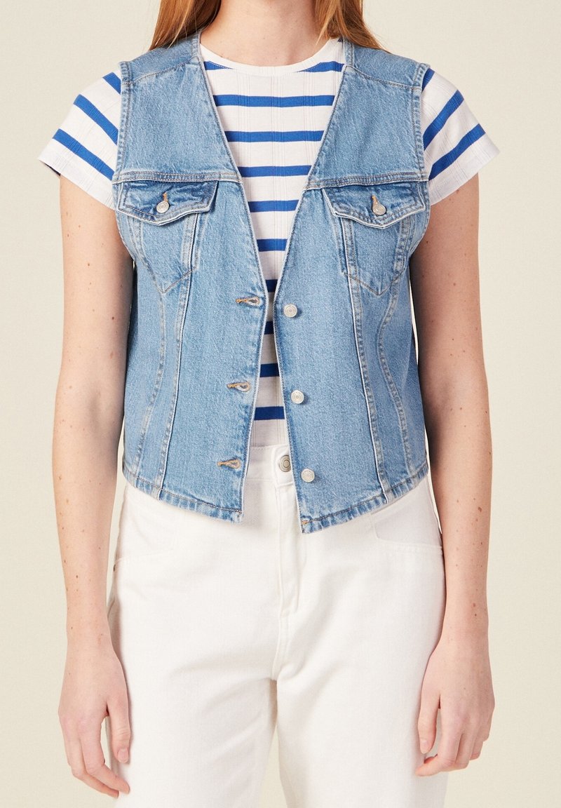 Jasnoniebieska denimowa kamizelka z dwiema kieszeniami na piersi, zapięciem na guziki i surowym wykończeniem. Nosi się ją na białej koszulce w niebieskie paski. Prosty design.