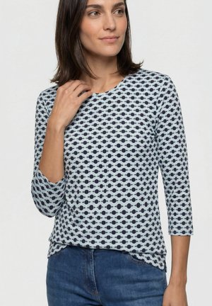 Frau trägt ein eng anliegendes, helles Oberteil mit marineblauem Kreis-Muster und Dreiviertelärmeln, kombiniert mit blauen Jeans.